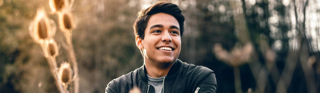 Dentist Canoga Park CA Dental Checkup Focused A young man smiles confidently while standing outdoors in nature, surrounded by dried plants and soft sunlight.