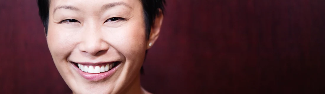 A close-up features a smiling person with short, dark hair, set against a blurred reddish-brown background.