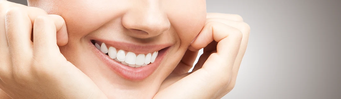A close-up captures a wide smile showcasing very white teeth while hands gently frame the sides of the smiling mouth.